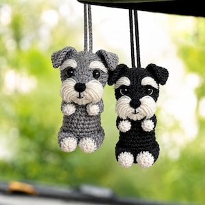 Pode incluir: Dois ornamentos de cão feitos em crochê pendurados por cordões. Um cão é cinzento com detalhes brancos no focinho e nas patas, enquanto o outro é preto com detalhes brancos semelhantes. Ambos têm olhos escuros e orelhas caídas em fio.