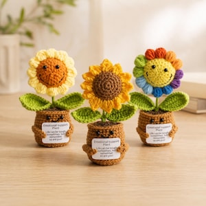 Könnte beinhalten: Drei gehäkelte Sonnenblumen in braunen Töpfen mit lächelnden Gesichtern. Jeder Topf hält ein weißes Schild mit der Aufschrift: Emotional support Plant. Life can be tough sometimes, but remember so are you! Blüten in Gelb, Orange und Regenbogenfarben.