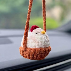 May include: A crocheted white chicken plush toy with a small red comb and yellow beak, nestled inside a brown crocheted basket. The basket is suspended by two long, braided brown yarn straps, designed to hang as a decorative ornament.