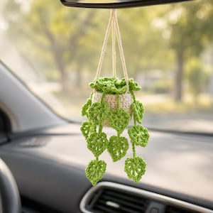 May include: A crocheted hanging plant ornament featuring a small, cream-colored pot with vibrant green, heart-shaped leaves trailing downwards. The plant is suspended by a cream-colored yarn cord, designed to hang from a vehicle rearview mirror.