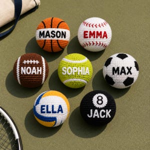 Puede incluir: Una colección de siete pelotas deportivas de ganchillo con nombres personalizados: MASON en un balón de baloncesto naranja, EMMA en una pelota de béisbol blanca, NOAH en un balón de fútbol americano, SOPHIA en una pelota de tenis amarilla, MAX en un balón de fútbol, ELLA en un balón de voleibol y JACK en una bola de billar negra número 8.