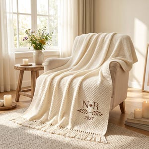 Può includere: Una coperta in maglia a nido d'ape color crema con bordi sfrangiati, drappeggiata su una poltrona. La coperta presenta un ricamo marrone con le lettere N e R separate da un cuore, un motivo a rametti e l'anno 2027 nella parte inferiore.