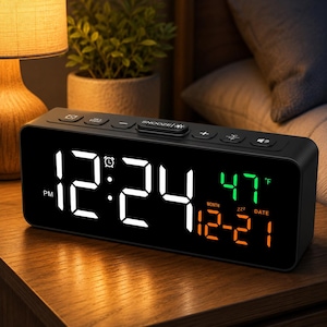 May include: A rectangular black digital alarm clock with a large white LED display showing 12:24 PM. The screen features a green temperature reading of 47 degrees Fahrenheit and an orange date display of 12-21. Buttons for snooze and settings are on top.