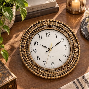 May include: A round wall clock with a textured gold-toned frame featuring a beaded border. The clock face is off-white with black Arabic numerals, black hour and minute hands, and a thin gold-toned second hand. The word Quartz is printed in black.