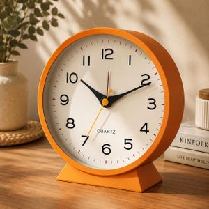 May include: A round orange quartz table clock with a white face, black Arabic numerals, and black hour and minute hands. A thin yellow second hand extends from the center. The clock features a sturdy orange base and the word QUARTZ printed below.