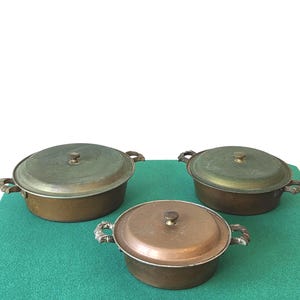 May include: Three round copper pots with matching lids and decorative metal side handles. The pots vary in size and feature a weathered, aged patina on the surfaces. Each lid is topped with a small, centered knob for easy lifting and handling.