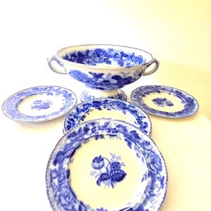 Könnte beinhalten: Eine weiße Keramik-Servierschale mit zwei Griffen und vier passenden Tellern, alle mit einem blauen Blumenmuster verziert. Die Schale hat einen Standfuß und jedes Teil weist einen gewellten Rand mit detaillierten blauen Blumenmotiven auf.