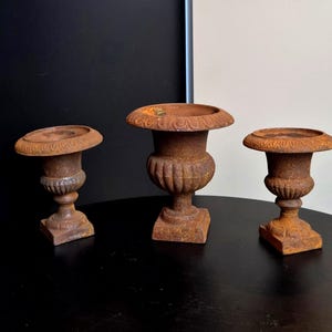 Op de afbeelding: Een set van drie gietijzeren urnen met een verweerde, roestbruine afwerking. Elke urn heeft een brede, ronde rand met decoratieve details, een bolvormig lichaam met verticale groeven, een slanke voet en een vierkante onderkant.