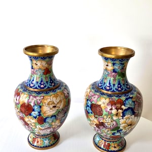 May include: A pair of cloisonné vases featuring a vibrant floral pattern on a light green background. The vases have a brass rim, neck, and base, with intricate enamel details in shades of red, blue, yellow, and purple depicting various flowers.