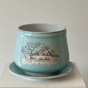 May include: A light blue ceramic planter with a matching saucer, featuring a brown ink illustration of a winter landscape with a house and bare trees on the front. The planter has a smooth, glossy finish and a simple, rounded cylindrical shape.