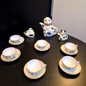 Op de afbeelding: Een keramieken theeservies bestaande uit een katvormige theepot met een oranje halsband, een bijpassend melkkannetje, een suikerpot met deksel en zes kopjes met schotels. Elk item is wit met zwarte vlekken en een dunne oranje rand langs de randen.