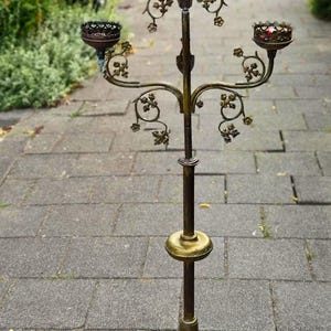 May include: A tall, multi-armed brass candelabra featuring five candle holders with crown-like rims. The central stem is adorned with decorative metal leaf motifs and branches. The base consists of a reddish stone section atop a wide, circular brass disc.