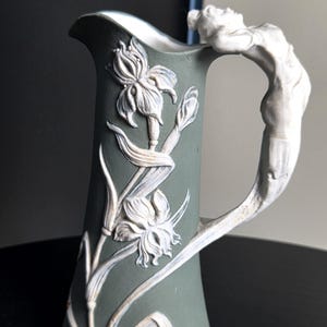 Può includere: Una brocca in ceramica verde salvia con disegni floreali di iris in rilievo bianco sul corpo e una figura bianca scolpita che forma il manico. La brocca presenta un bordo svasato e una finitura opaca liscia con dettagli botanici.