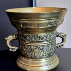 May include: A brass mortar featuring a wide, flared rim with engraved letters and numbers, including 3, Z, and LO. The body displays intricate relief patterns of scrolling foliage and two side handles shaped like stylized animal heads.