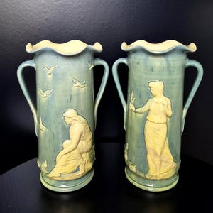 Peut inclure: Deux vases en céramique avec un vernis bleu clair et des figures en relief de couleur crème. Chaque vase présente deux anses, un bord ondulé et des motifs en relief représentant une personne et des oiseaux. Les figures portent des vêtements fluides.