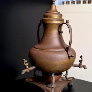 Op de afbeelding: Een koperkleurige metalen samovar met een centrale ketel inclusief deksel, twee zijhandvatten en twee kranen aan de onderzijde. Het geheel rust op een driepotige standaard met een klein kommetje eronder en heeft een verweerde patina afwerking.