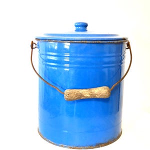 Pode incluir: Um recipiente cilíndrico de metal esmaltado azul com tampa a condizer. Possui uma pega giratória de arame enferrujado com um punho central de madeira. A base e o rebordo apresentam sinais de oxidação e desgaste no metal.