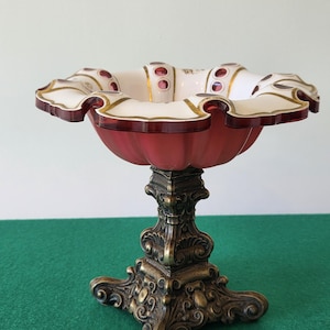 Può includere: Una coppa decorativa su piedistallo con bordo smerlato, accenti in vetro rosso scuro e pannelli bianchi. La coppa è sostenuta da una base in metallo decorata color bronzo scuro con volute intricate e un design quadrato a quattro piedi.