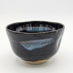 May include: A small, round ceramic bowl with a glossy, dark black glaze. The rim and upper interior feature a subtle, mottled blue and white drip pattern. The base of the bowl is unglazed, revealing a light tan, matte clay texture.