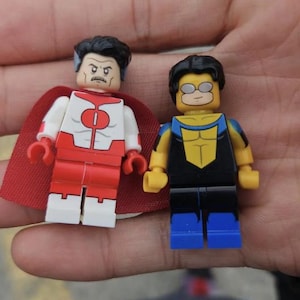 May include: Two Lego minifigures of comic book characters. One figure has a red and white suit with a red cape. The other figure has a black and yellow suit with blue boots and yellow arms. Both figures have black hair.