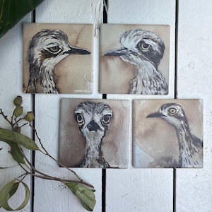 May include: Four square ceramic coasters featuring watercolour-style illustrations of stone-curlew birds in shades of brown, grey, and white. Each coaster displays a unique bird portrait with the artist name Hannah Mello printed in the bottom corner.