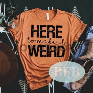 May include: A burnt orange t-shirt with black text that reads "HERE to make it WEIRD". The shirt is laid out on a black and white patterned surface with a brown hat and blue jeans.