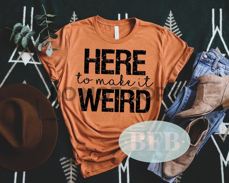 May include: A burnt orange t-shirt with black text that reads "HERE to make it WEIRD". The shirt is laid out on a black and white patterned surface with a brown hat and blue jeans.