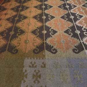 May include: Close-up of a patterned rug with a geometric design. The rug features a repeating pattern of diamonds and other shapes in shades of brown, orange, and gray. The rug has a textured appearance.
