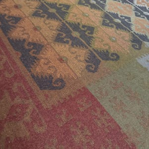 May include: Close-up of a patterned rug with a geometric design. The rug features a color palette of red, brown, orange, green, and black. The design includes repeating shapes and patterns, creating a textured appearance.