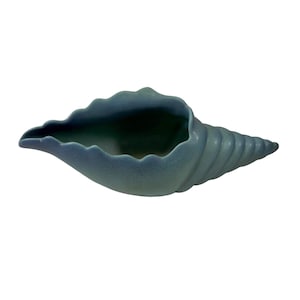 Puede incluir: Un cuenco decorativo de cerámica azul con forma de concha. El cuenco tiene una superficie texturizada y una amplia abertura con un borde festoneado. El diseño en espiral se estrecha hasta un punto. El cuenco es de color azul apagado.