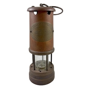 May include: Vintage brown metal miner's lamp with a clear glass enclosure. The lamp has a handle and a brass plate with text that reads "British Coal Mines". The lamp is cylindrical and has a round base.