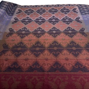 May include: A folded blanket with a geometric pattern in shades of blue, peach, brown, and burgundy. The design features repeating diamond and zigzag shapes, creating a textured appearance. The blanket is likely made of a soft material.