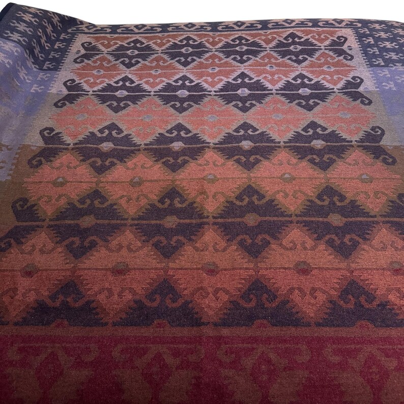 May include: A folded blanket with a geometric pattern in shades of blue, peach, brown, and burgundy. The design features repeating diamond and zigzag shapes, creating a textured appearance. The blanket is likely made of a soft material.