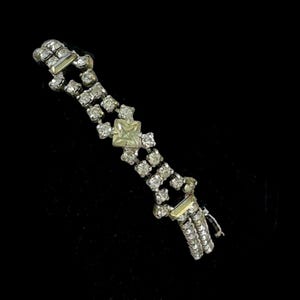 May include: A silver-toned bracelet with multiple rows of small, clear rhinestones. A square, pale yellow gemstone is centered on the bracelet. The bracelet is set against a black background.