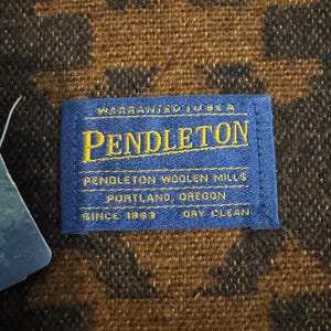 May include: Close-up of a blue Pendleton label with gold lettering on a brown and black patterned fabric. The label reads "Warranted to be a Pendleton" and "Pendleton Woolen Mills, Portland, Oregon, Since 1863, Dry Clean."