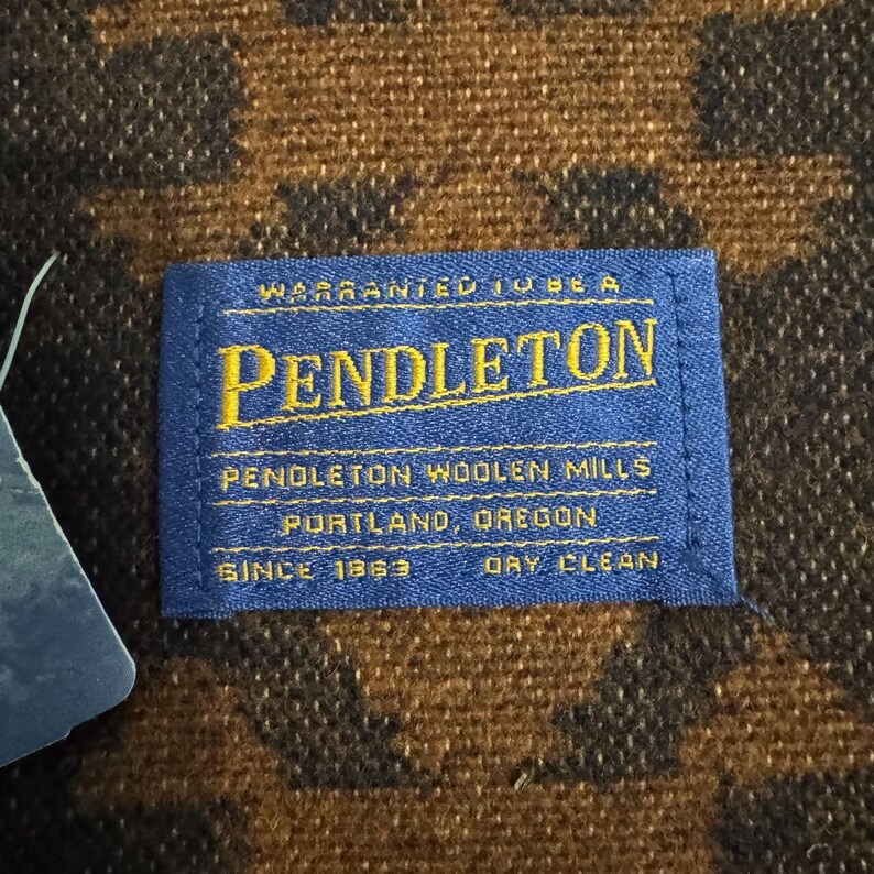 May include: Close-up of a blue Pendleton label with gold lettering on a brown and black patterned fabric. The label reads "Warranted to be a Pendleton" and "Pendleton Woolen Mills, Portland, Oregon, Since 1863, Dry Clean."