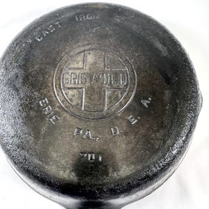 May include: Close-up of a cast iron skillet base. The skillet is dark grey with the embossed text "GRISWOLD ERIE PA. U.S.A." and the number "701". The word "IRON" is also embossed on the skillet.