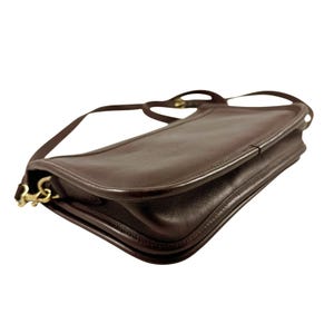 K&ouml;nnte beinhalten: Eine dunkelbraune Lederschultertasche mit glattem, abgerundetem Klappenverschluss und einem d&uuml;nnen, passenden Lederriemen. Die Tasche verf&uuml;gt &uuml;ber goldfarbene Metallbeschl&auml;ge, einschlie&szlig;lich eines Drehverschlusses an der Seite, und eine strukturierte Form.