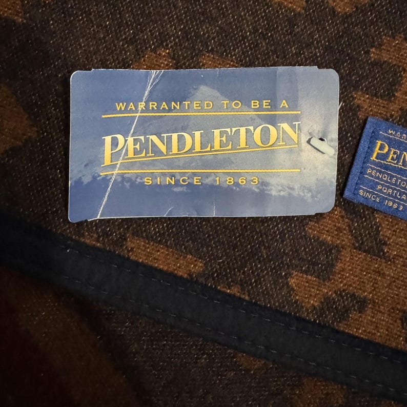 May include: Close-up of two blue and white Pendleton tags. The larger tag reads "WARRANTED TO BE A PENDLETON SINCE 1863" in gold lettering. The background is a brown and black patterned fabric with a black trim.
