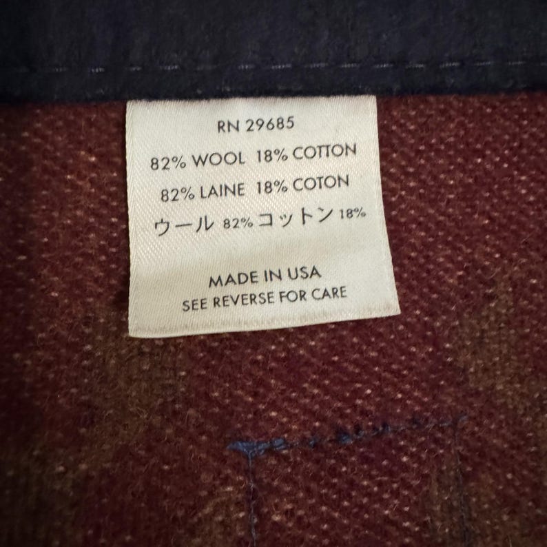 May include: Close-up of a white tag with black text, showing fabric composition and care instructions. The tag reads "RN 29685" and lists the materials as 82% wool and 18% cotton. The tag also states "Made in USA."
