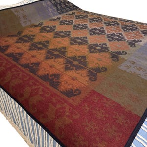 May include: A colorful blanket with a geometric pattern in shades of orange, brown, and burgundy. The blanket has a fringed edge and a dark blue border. The design features repeating diamond shapes and floral motifs.