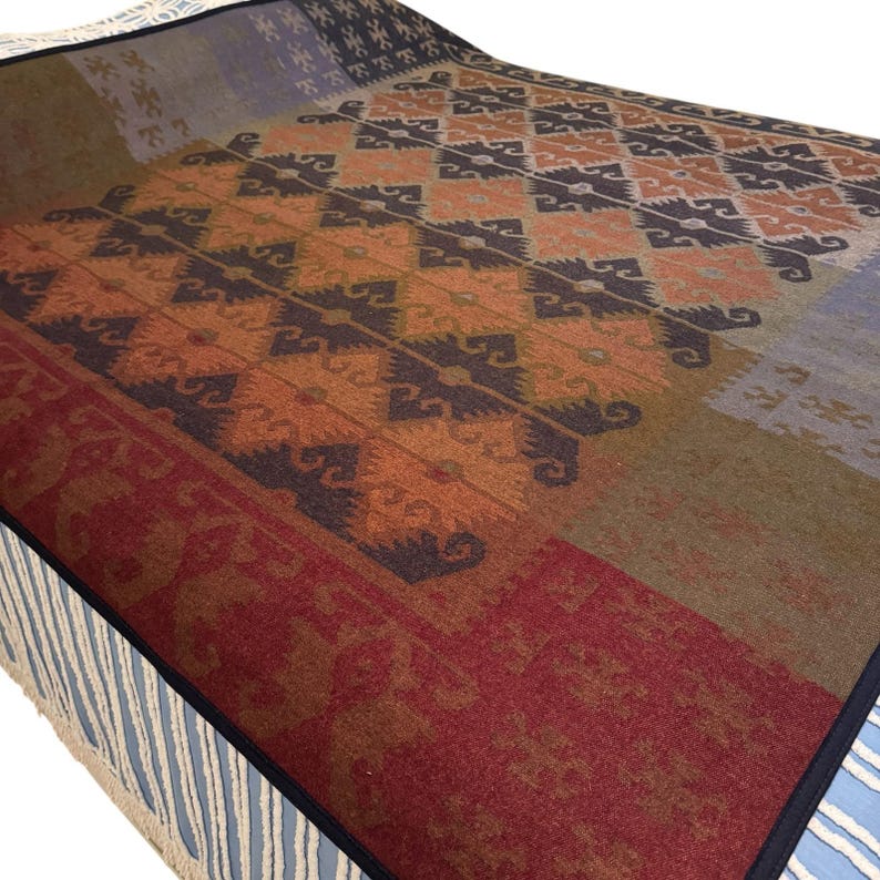 May include: A colorful blanket with a geometric pattern in shades of orange, brown, and burgundy. The blanket has a fringed edge and a dark blue border. The design features repeating diamond shapes and floral motifs.