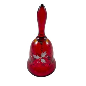 May include: A vibrant ruby red glass bell with a long handle. The bell has a white etched design of leaves and berries. This decorative bell is likely for display or collecting.