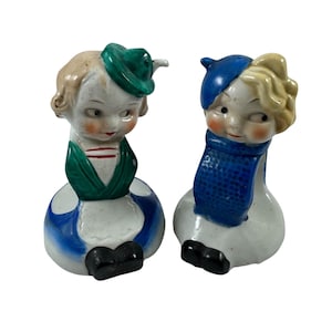 May include: A pair of vintage ceramic figurines, each depicting a child's head and torso. One wears a green hat and vest, the other a blue hat and scarf. Both have rosy cheeks and are set on a white base with blue and black accents.