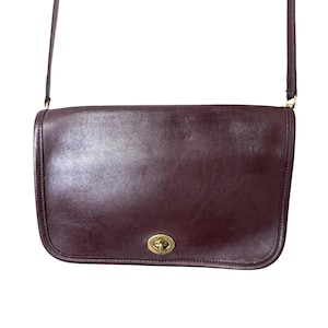K&ouml;nnte beinhalten: Eine dunkelburgunderfarbene Lederschultertasche mit glatter Oberfl&auml;che und rechteckiger Silhouette. Die Tasche verf&uuml;gt &uuml;ber einen goldfarbenen Metalldrehverschluss, der mittig auf der Frontklappe sitzt, sowie einen d&uuml;nnen, passenden Schulterriemen.