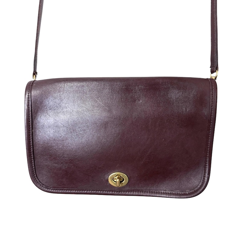 K&ouml;nnte beinhalten: Eine dunkelburgunderfarbene Lederschultertasche mit glatter Oberfl&auml;che und rechteckiger Silhouette. Die Tasche verf&uuml;gt &uuml;ber einen goldfarbenen Metalldrehverschluss, der mittig auf der Frontklappe sitzt, sowie einen d&uuml;nnen, passenden Schulterriemen.
