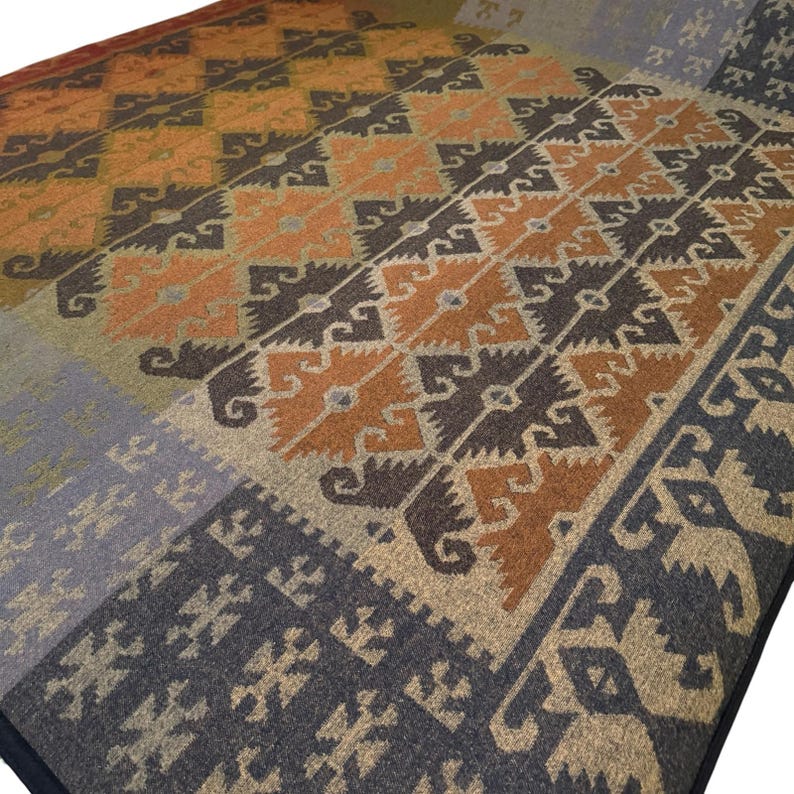 May include: A woven area rug with a geometric pattern in shades of orange, brown, gray, and beige. The rug features a repeating design of stylized shapes and a border in a darker gray. The rug is folded over.