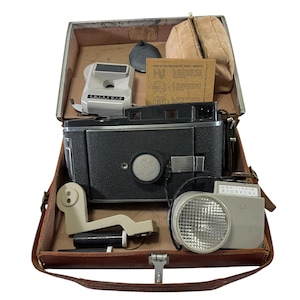 May include: Vintage Polaroid camera kit in a brown leather case. The kit includes a black camera, flash, and accessories. The case is open, revealing the contents. A beige pouch and instruction card are also visible.