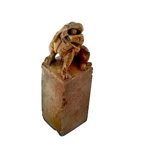Puede incluir: Una escultura de piedra marrón tallada de una criatura mítica sobre una base rectangular. La criatura tiene una apariencia de león con detalles precisos. La escultura es probablemente un artículo antiguo o vintage, que muestra una artesanía intrincada y una apariencia desgastada.