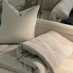 May include: A beige linen-textured throw blanket folded over a matching beige sofa cushion. The blanket features a subtle woven pattern and a soft, matte finish. A large, square, solid beige throw pillow rests upright against the back of the sofa.